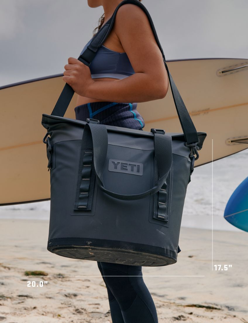 Person holding a YETI cooler bag with a surfboard in the background on a beach.