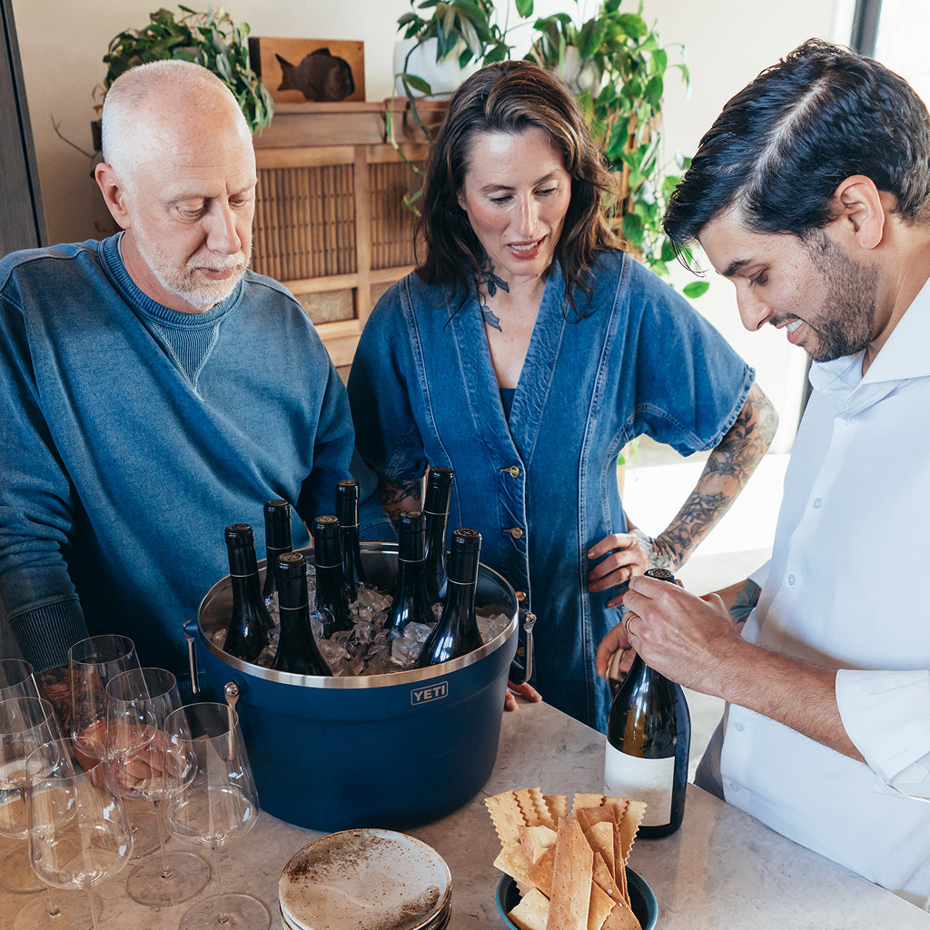 Three people around a table with wine bottles and glasses, one person opening a bottle.