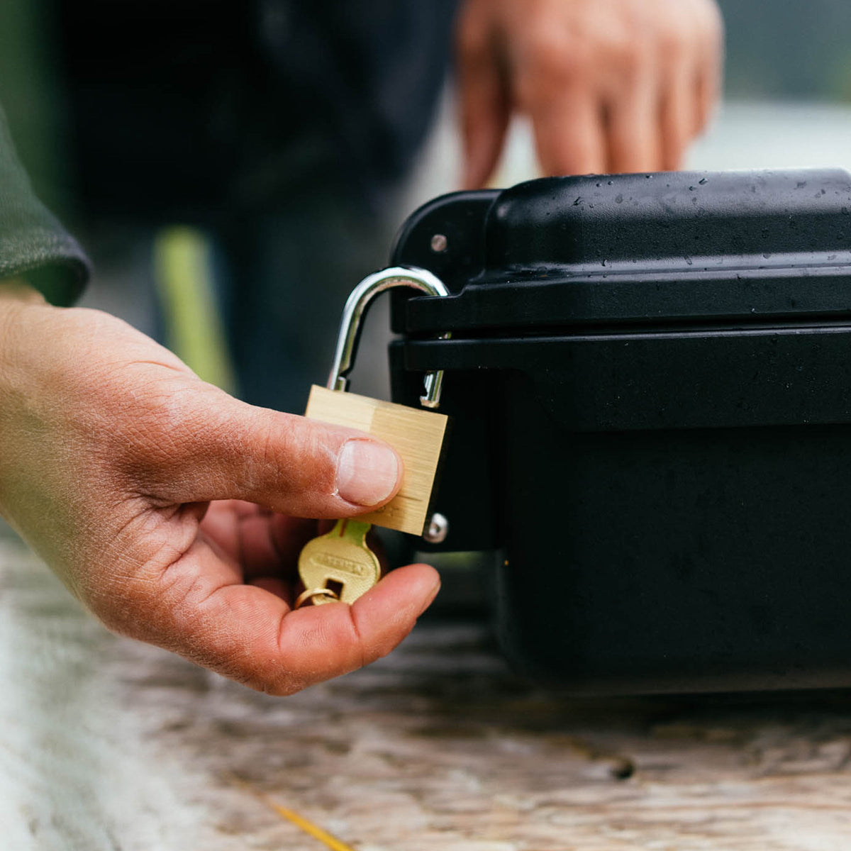 A close-up of a hand holding a brass padlock and key next to a black hard case, with another person's hand blurred in the background.