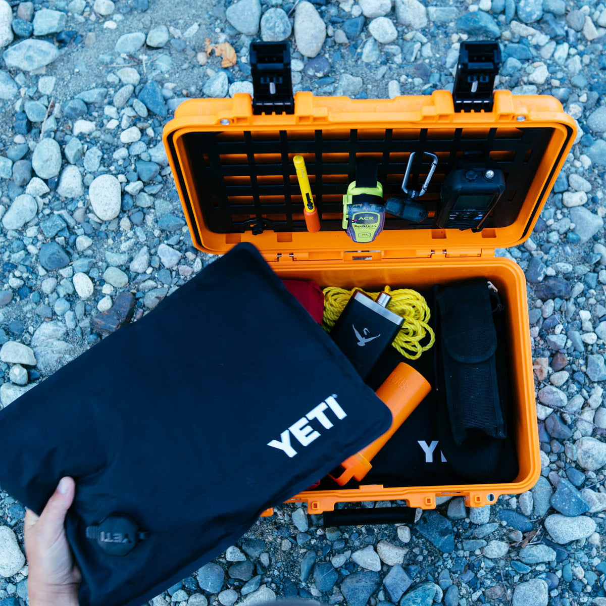 An open bright orange YETI hard case on a bed of gray pebbles, with a hand holding a black YETI pouch in front. Inside the case are various outdoor gear items including a yellow cord, an orange bottle, and other accessories.