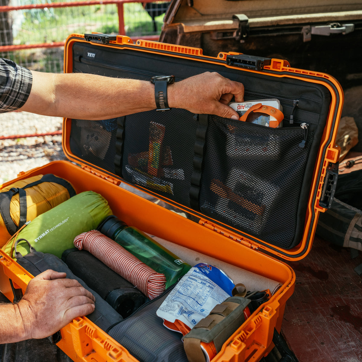An open bright orange hard-shell carrying case with mesh pockets and organizer compartments, shown being packed with rolled sleeping bags, a green sleeping pad, and other outdoor gear. A person's hands are visible organizing the contents inside.