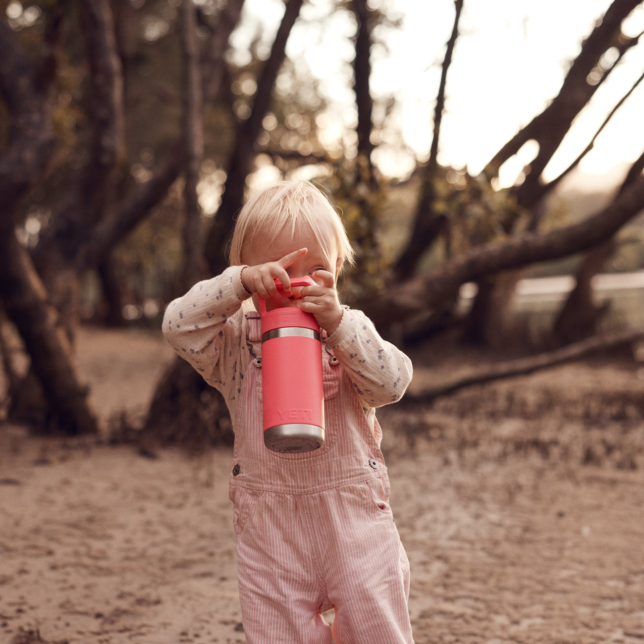 Child holding a pink YETI bottle in a natural setting with trees and water.
