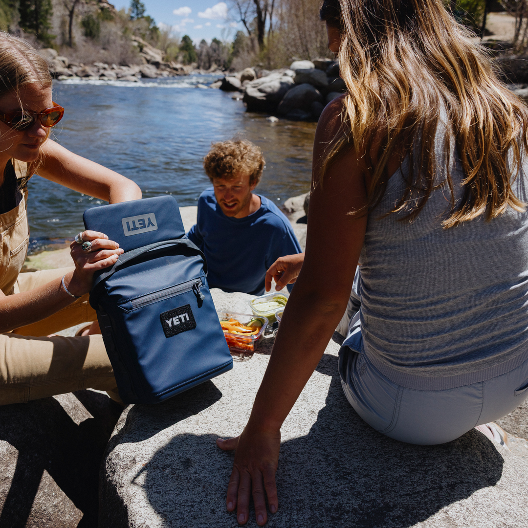 Two people interacting with a YETI cooler by a body of water.