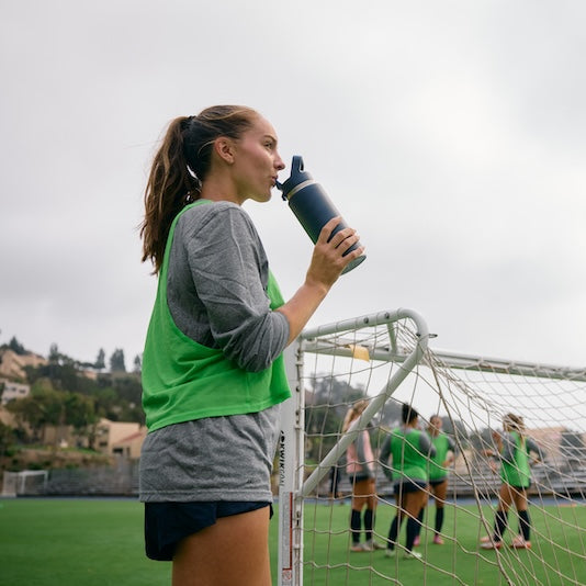 Woman drinking from a YETI water bottle on a soccer field with other players in the background.
